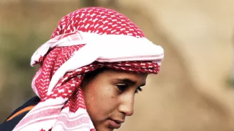 در این تصویر، یک فرد با چهرهای متعلق به سن جوانی دیده میشود. او یک کلاهی با طرح قرمز و سفید بر سر دارد که به شیوهای سنتی پوشیده شده است. چهرهاش به سمت پایین متمایل است و نگاهی تفکرآمیز دارد. پسزمینه تصویر مبهم است و توجه بیشتر بر روی شخص متمرکز شده است. این تصویر حس آرامش و تمرکز را منتقل میکند.