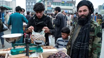 در تصویر، یک بازار شلوغ دیده میشود که مردانی و کودکانی در آن مشغول خرید و فروش هستند. در پیشزمینه، یک مرد با ریش بلند و لباسی سنتی در کنار یک میز است که بر روی آن مجموعهای از خرماها قرار دارد. او به نظر میرسد که در حال وزن کردن خرماها است. در سمت راست، مرد دیگری هم با لباسی مشابه و کلاهی بر سر ایستاده و به دور و برش نگاه میکند. جمعیتی از افراد دیگر نیز در پسزمینه دیده میشوند و حس شلوغی و فعالیت در بازار به خوبی منتقل شده است.