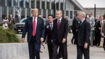 در این تصویر، چند مرد در حال پیادهروی هستند. یکی از آنها که لباس رسمی پوشیده و کلاهی تیره به سر دارد، به سمت دیگران نگاه میکند. شخص دیگری که در سمت چپ او قرار دارد، کت و شلوار و یک کراوات قرمز به تن دارد. دو مرد دیگر نیز به آنها ملحق شدهاند و ظاهراً در حال گفتگو هستند. پسزمینه، شامل ساختمانهای مدرن و تعدادی از افراد دیگر در حال حرکت است. جو کلی تصویر نشاندهنده یک ملاقات رسمی است.