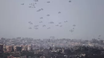 در این تصویر، چندین چتر نجات در آسمان به طور همزمان دیده میشوند که به آرامی در حال سقوط هستند. زمینه تصویر شهری است که شامل ساختمانها و محلههای مختلف است. آسمان ابری و کمی تاریک به نظر میرسد و این باعث میشود فضای تصویر حالتی جدی و نگرانکننده داشته باشد. تصویر به خوبی نمایانگر یک وضعیت نظامی یا اضطراری است.