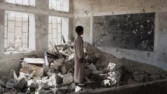 در این تصویر، یک پسر جوان در یک کلاس درس ویران شده ایستاده است. دیوارها و سقف کلاس آسیب دیدهاند و بخشهایی از زیرساختها به طور کامل تخریب شدهاند. در اطراف او، زبالهها و آوارهای ساختمانی وجود دارد. یک تابلو سیاه با نوشتههایی بر روی دیوار وجود دارد، اما وضعیت آن نیز نامناسب است. نور از پنجرههای شکسته به فضای داخلی میتابد و حس ناامیدی و ویرانی را منتقل میکند.