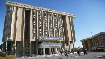 این تصویر نمای ساختمان پارلمان کردستان در عراق را نشان میدهد. ساختمان دارای طراحی مدرن و بزرگ است و چندین طبقه دارد. در جلوی ساختمان، پرچمهای کردستان و عراق به اهتزاز درآمدهاند. همچنین، افرادی در حال تردد در محوطه مقابل ساختمان دیده میشوند. آسمان آبی و صاف به زیبایی محیط افزوده است.