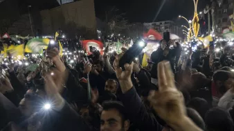 در این تصویر، جمعیتی پرشور و هیجانزده دیده میشود که در یک شب تاریک گرد هم آمدهاند. افراد حاضر در مراسم، پرچمهایی با رنگهای مختلف را در دست دارند و برخی از آنها نیز با استفاده از گوشیهای خود چراغ قوهها را روشن کردهاند. فضای عمومی شلوغ و مملو از شور و احساسات اجتماعی یا سیاسی به نظر میرسد. جماعت به سمت دوربین یا نقطهای خاص معطوف شده و برخی با دست خود علامتهایی را نشان میدهند.