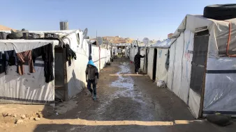 این تصویر نمایی از یک کمپ پناهندگان را نشان میدهد. در اینجا، چادرهای سفید و مشکی به صورت ردیفی در دو طرف یک مسیر خاکی قرار دارند. زمین به نظر میرسد کمی مرطوب و گلآلود است. چندین قطعه لباس بر روی چادرها آویزان شدهاند و شخصی در حال حرکت در مسیر دیده میشود. در پسزمینه، ساختمانهایی در بالای تپهها قابل مشاهده هستند. این منظره فضایی ساده و دلگیر را تداعی میکند.