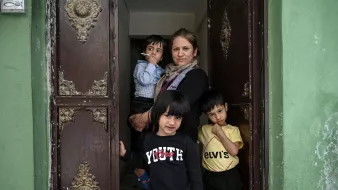 در این تصویر، یک زن با موهای قهوهای و لباس گرم در ورودی یک ساختمان ایستاده است. او یک سیگار در دست دارد. در کنار او دو کودک کوچک و یک کودک دیگر در آغوش او هستند. یکی از کودکان در پیشزمینه با لباسی تیره و نوشتهای روی آن ایستاده و به دوربین نگاه میکند. دیگر کودکان نیز در پشت او ایستادهاند و به نظر میرسد که در حال صحبت یا بازی هستند. دیوارهای اطراف ورودی ساختمان به رنگ سبز و با جزئیات قدیمی تزئین شدهاند. فضای تصویر صمیمی و خانوادگی است.