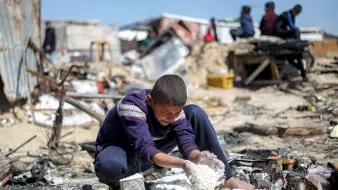 پسرکی در میان خرابهها نشسته و مشغول بازی با مواد سفید رنگ است. فضای اطراف بسیار بینظم و ویران است.