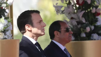 این تصویر دو مرد را در کنار هم نشان میدهد. شخصی در سمت چپ با موهای تیره و لباس رسمی مشکی، در حالت تفکر و جدی ایستاده است. در سمت راست، مرد دیگری با عینک آفتابی و لباس رسمی به رنگ تیره، در کنار او قرار دارد. پسزمینه شامل گلهای سفید و عناصر طبیعی است که به تصویر زیبایی و شکیل بودن میبخشد.