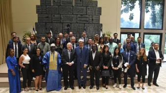 این تصویر گروهی از افراد را نشان میدهد که در یک مکان رسمی و با پسزمینهای زیبا جمع شدهاند. در وسط تصویر، چند مرد در کت و شلوار ایستادهاند که به نظر میرسد مسئولین یا مقامهای بالاتر باشند. بقیه افراد، شامل مردان و زنانی با لباسهای متنوع، در اطراف آنها ایستادهاند و همگی به دوربین نگاه میکنند. در پسزمینه، پرچمها و یک دیوار زیبا مشاهده میشود که به محیط رسمی فضا افزوده است. افراد در این عکس به نظر میرسد که از ملیتها و فرهنگهای مختلفی هستند.