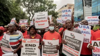 در این تصویر، گروهی از معترضان در حال برگزاری تجمعی هستند. آنها پوسترهایی با شعارهای ضد اسرائیلی و حمایتی از فلسطین را در دست دارند. برخی از شعارها شامل «تحریم اسرائیل» و «پایان آپارتاید اسرائیل» هستند. معترضان با لباسهای مشکی و قرمز دیده میشوند و به نظر میرسد که به شدت بر علیه سیاستهای اسرائیل و در حمایت از حقوق فلسطینیان اعتراض میکنند. پسزمینه تصویر شامل ساختمانهای اداری و درختان است.