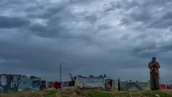 در این تصویر، یک مرد در منطقهای با ساختمانهای موقتی یا کمپ زندگی میکند. آسمان ابری و تیره به نظر میرسد و نشاندهنده شرایط جوی نامساعد است. در پسزمینه، چند چادر یا خانه موقت دیده میشود که با پارچهها و وسایل مختلف پوشانده شدهاند. فضای کلی تصویر حس کمبود و در عین حال استقامت را منتقل میکند. مرد به نظر میرسد در حال مکالمه یا تفکر است و تصویر کلی فضایی جدی و غمانگیز دارد.
