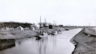 تصویر نمایی قدیمی از یک رودخانه یا کانال است که در دو طرف آن قایقها، به ویژه قایقهای بادبانی، قرار دارند. در سمت راست تصویر، چندین قایق به آرامی در آب شناور هستند و در سمت چپ، بخشی از ساحل با شن و ماسه دیده میشود. در پسزمینه، چادرها و ساختمانهایی به چشم میخورد که نشاندهنده حضور انسانی و فعالیتهای مربوط به کشاورزی یا صید هستند. نور تصویر به نظر میرسد نرم و ملایم باشد که حس آرامشی را ایجاد میکند.