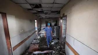 مردی با کلاه ایمنی در راهروی ویران یک بیمارستان قدم میزند، دیوارها آسیب دیده و کف پر از آوار است.