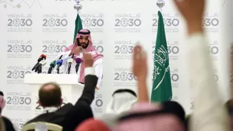 در این تصویر، یک مرد جوان با لباس سنتی سعودی و عمامه، در حال سخنرانی در مقابل میکروفنها و دوربینهای خبری است. او در پسزمینهای شامل پرچمهای عربستان سعودی و شعارهایی درباره "چشمانداز ۲۰۳۰" قرار دارد. تعدادی از افراد نشسته در سالن، دستهای خود را برای پرسش بالا بردهاند. فضا به وضوح نشاندهنده یک کنفرانس خبری یا رویداد رسمی است.