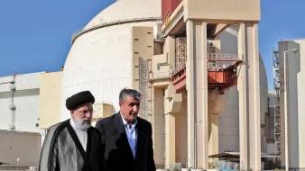 در این تصویر، دو فرد در حال قدم زدن در کنار یک ساختمان بزرگ هستند که به نظر میرسد یک نیروگاه هستهای باشد. ساختمان دارای ديواری گرد و بزرگ با رنگ روشن و نوارهایی است که به طور مشخص بر روی آن قرار دارد. آسمان در پسزمینه آبی و روشن است. افراد حاضر در تصویر لباسهای رسمی به تن دارند. یکی از آنها عمامهای بر سر دارد و ظاهراً مقام بالایی در نظام دارد. تصویر به نوعی به رویدادهای مرتبط با انرژی هستهای اشاره دارد.