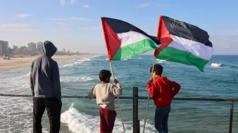 در این تصویر، سه کودک در کنار دریا ایستادهاند و به سمت امواج دریا نگاه میکنند. یکی از کودکان پرچم فلسطین را در دست دارد و دو نفر دیگر نیز در حال نظارت بر صحنه هستند. لباسهای آنها متنوع است و در پسزمینه میتوان نمایی از سواحل و ساختمانهای شهری را مشاهده کرد. آسمان آبی و ابرهای کمی در بالای تصویر وجود دارد که حال و هوای دلپذیری را به تصویر میدهد.