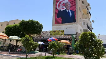 در این تصویر، یک دیوار بزرگ ساختمان نشان داده شده است که بر روی آن تصویری از یک فرد با اشاره به سمت بیننده مشاهده میشود. این شخص لباس رسمی به تن دارد و در پسزمینه تصویر، پرچم تونس دیده میشود. در پایین تصویر، یک کارگاه یا فروشگاه کوچک قرار دارد که چند نفر در زیر چادرهای رنگی نشستهاند. درختان سبز و دوچرخهها نیز در تصویر وجود دارند. آسمان آبی و صاف به نظر میرسد.