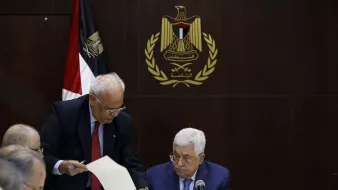 در این تصویر، یک جلسه رسمی در حال برگزاری است. در مرکز تصویر، مردی به نام محمود عباس نشسته است که به نظر میرسد در حال مطالعه مدارک یا مستندات است. افراد دیگری نیز در اطراف او نشستهاند. یکی از آنها به سمت عباس خم شده و به او مدرکی نشان میدهد. در پسزمینه، پرچم فلسطین و نماد رسمی مرتبط با دولت فلسطین دیده میشود، که به فضایی رسمی و سیاسی اهمیت میدهد. میز گردی که آنها دور آن نشستهاند، با لوازم اداری و بطریهای آب تزیین شده است.