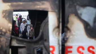 تصویر نشاندهندهی یک صحنهی غمانگیز است که در آن چندین کودک و بزرگسال درحال مشاهدهی یک وضعیت بحرانی به نظر میرسند. در پسزمینه، یک وسیله نقلیه آسیبدیده دیده میشود که حالت سوخته و خراب دارد. نام "پرس" بر روی آن به وضوح مشاهده میشود. در داخل نمای این وسیله، افرادی با حالتهایی جدی و نگران دیده میشوند که شاید به دنبال کمک یا راهی برای فرار از وضعیت خطرناک باشند. احساس تنش و اضطراب در این تصویر محسوس است.