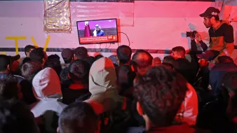 در تصویر، گروهی از افراد در حال تماشای تلویزیونی هستند که بر روی دیواری نصب شده است. تلویزیون یک برنامه خبری را نمایش میدهد و یک سخنران در حال صحبت است. افراد به سمت صفحه تلویزیون خیره شدهاند و جو عمومی تصویر نشاندهنده توجه و کنجکاوی آنهاست. فضای اطراف کمی تاریک و نور محیطی کم است، به طوری که نور تلویزیون بیشتر مشخص است. برخی از افراد در لباسهای گرم، نظیر هودی و کلاه، حضور دارند. یکی از افراد در سمت راست با دوربین در حال عکاسی است.