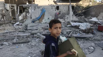 این تصویر یک پسر جوان را نشان میدهد که در میان ویرانههای یک ساختمان آسیبدیده ایستاده است. او در حال حمل یک جعبه است و به سمت دوربین نگاه میکند. محیط اطراف او پر از زباله، آوار و تکههای مصالح ساختمانی است. دیوارهای خراب و نشانههای تخریب در پسزمینه مشهود است و وضعیت کلی تصویر نشاندهنده یک منطقه آسیبدیده از جنگ یا بلایای طبیعی است. احساساتی نظیر ناامیدی و تلخی در چهره کودک منعکس شده است.