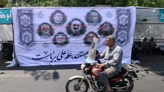 تصویر مردی سوار بر موتورسیکلت که در پیشزمینه، بنر بزرگ و مردم نشسته در پشت آن دیده میشوند.