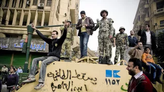 در این تصویر، یک گروه از مردان و سربازان در حال تجمع روی یک خودرو نظامی هستند. چند نفر از آنها به سمت دوربین خوشحالی میکنند و دست بلند کردهاند. در پسزمینه، ساختمانهایی با نشانهها و نوشتههایی به زبان عربی دیده میشود. فضای تصویر نشاندهنده یک وضعیت اجتماعی یا سیاسی است که در آن افراد در حال ابراز احساسات و تعامل با یکدیگر هستند.