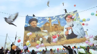 در این تصویر، تعدادی از مردم در حال برگزاری یک مراسم یا جشن هستند. در پسزمینه، یک دیوار بزرگ با تصویری از دو شخصیت سیاسی یا مذهبی به نمایش درآمده است. افراد در حال پرتاب بالنهای رنگی و همچنین برخی گلها به سمت هوا هستند. برخی از مردم در حال رقص و شادمانی دیده میشوند. پرندههایی نیز در آسمان در حال پرواز هستند و فضای کلی تصویر روحیه مثبت و شادابی را به نمایش میگذارد.