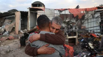 در این تصویر، دو مرد به یکدیگر در آغوش میافتند. پسزمینه تصویر نشاندهنده یک محل ویران شده است که به نظر میرسد دچار آتشسوزی یا تخریب شده است. حس ناراحتی و همدردی در چهرههای آنها مشهود است و فضای کلی تصویر احساساتی و غمانگیز به نظر میرسد.