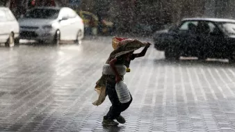 در این تصویر، کودکی در حال دویدن زیر باران شدید است. او یک کیسه یا پارچهای به سر دارد که به عنوان محافظ استفاده میکند و در دستش یک ظرف آب یا شیر میگیرد. قطرات باران به شدت به زمین میریزند و محیط اطرافش را همراه با نور و سایههای مختلف تحت تأثیر قرار دادهاند. خیابان کمی خیس است و در پسزمینه، خودروهایی دیده میشوند که در حال حرکت هستند. جوّ تصویر حس همدلی و مبارزه با شرایط نامساعد را منتقل میکند.