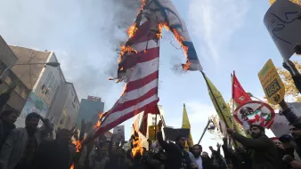 در این تصویر، جمعیتی در حال برگزاری یک تظاهرات هستند. گروهی از افراد در حال آتش زدن پرچمهایی با نمادهای مشخص هستند. پشت زمینه تظاهرات، ساختمانها و تابلوهای تبلیغاتی دیده میشود. شمار زیادی از تظاهرکنندگان در دست خود پلاکاردهایی نشان میدهند که بر روی آنها شعارهایی نوشته شده است. جو عمومی تصویر نشاندهندهٔ هیجان و احساسات قوی شرکتکنندگان است.