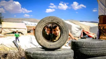 در این تصویر، دو کودک در حال نگاه کردن از میان یک لاستیک بزرگ هستند. لاستیکها به صورت عمومی به عنوان بخشی از یک ساخت و ساز یا بازی در محوطهای از فضای باز قرار داده شدهاند. پسزمینه شامل چادرها و مناظر طبیعی است. آسمان آبی و ابرهای پراکنده در بالای سر دیده میشوند. فضای کلی تصویر حس کنجکاوی و بازی کودکانه را منتقل میکند.