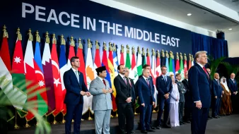 مردان در یک صف ایستادهاند، پرچمهای کشورهای مختلف پشت سرشان و نوشتهای بزرگ درباره "صلح در خاورمیانه" بالای سر.