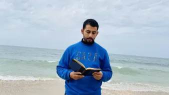 در این تصویر، مردی در کنار دریا ایستاده است و یک کتاب در دست دارد. او لباسی آبی رنگ به تن دارد که روی آن کلمه "GAZA" به چشم میخورد. پسزمینه تصویر را دریای آرام و آسمانی ابری تشکیل میدهد. موجهای دریا به ساحل میرسند و فضای آرامی را به تصویر بخشیدهاند.