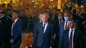 در این تصویر، گروهی از افراد در یک محیط شلوغ و پرجمعیت بهنظر میرسند. در مرکز تصویر، فردی با کت و شلوار تیره در حال عبور است و چندین محافظ یا همراه در اطراف او حضور دارند. برخی از افراد در زمینه تصویر، ماسک بهصورت دارند و دیگران مشغول تماشا و عکاسی از صحنه هستند. محیط به نظر میرسد شبانه باشد و نورها بهخوبی در تصویر مشاهده میشوند.