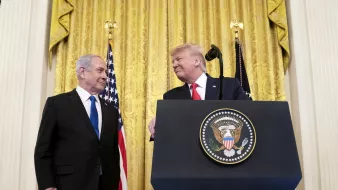 تصویر مربوط به یک مراسم رسمی است که در آن دو نفر در حال گفتگو هستند. در پسزمینه، پردههای طلایی و پرچمهای ایالات متحده دیده میشوند. یکی از افراد در سمت چپ، لباسی تیره به تن دارد و به شخص دیگر در سمت راست نگاه میکند. شخص در سمت راست یک لباس رسمی و یک کراوات به تن دارد و به نظر میرسد در حال لبخند زدن است. همچنین بر روی میزی که در مقابل آنها قرار دارد، نشان رسمی ریاستجمهوری ایالات متحده وجود دارد. این تصویر به نظر میرسد به یک رویداد سیاسی یا دیپلماتیک مربوط باشد.