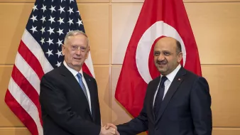 در این تصویر، دو مرد در حال دست دادن هستند. در پسزمینه، پرچمهای ایالات متحده و ترکیه دیده میشوند. یکی از مردان، با موهای خاکستری و کت و شلوار تیره، به نظر میرسد یک مقام آمریکایی باشد. مرد دیگر نیز با کت و شلوار تیره و کراواتی با طرحی خاص، نماینده ترکیه به نظر میرسد. این تصویر به نوعی نشاندهنده همکاری یا مذاکرات بین دو کشور است.