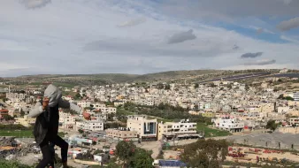 در این تصویر، فردی در حال بالا رفتن یا حرکت در مکانی کوهستانی دیده میشود. او لباسهای غیررسمی به تن دارد و در پسزمینه، یک شهر بزرگ با ساختمانهای مختلف و معماری متنوع دیده میشود. آسمان ابری است و مناظر طبیعی اطراف، مانند تپهها و درختان، نیز در تصویر قابل مشاهدهاند. این صحنه ترکیبی از زندگی شهری و طبیعت را نشان میدهد.