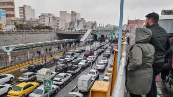 تصویر نشاندهنده یک صحنه شلوغ از ترافیک در یک خیابان شهری است. در سمت چپ تصویر، افرادی در حال تماشا از بالا ایستادهاند. در پایین، خطوط خودروها به وضوح قابل مشاهده است که نشاندهنده تجمع و ترافیک سنگین است. بقیه عناصر شهری مانند ساختمانها و علائم راهنمایی نیز در پسزمینه دیده میشوند. هوای ابری و احتمالا بارانی به دید فضا و حس و حال تصویر اضافه میکند.