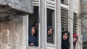 در این تصویر، تعدادی زن از پنجرههای یک ساختمان قدیمی و دارای نمایی خراب نگاه میکنند. آنها در حالتهای مختلفی قرار دارند و به نظر میرسد نگران یا کنجکاو هستند. پوشش آنها شامل چادر و روسری است که نشاندهنده فرهنگ خاصی است. فضای تصویر به طور کلی تلخ و پر از تنشهای اجتماعی به نظر میرسد.