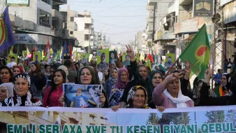 تصویر یک تظاهرات بزرگ را نشان میدهد که گروهی از زنان در آن شرکت دارند. آنها در خیابانی شلوغ راه میروند و پلاکاردهایی به دست دارند. تعدادی پرچم با نمادهایی خاص نیز در دست دارند. زنان با لبخند و دستهای بالا به نشانه حمایت و ابراز احساسات مشارکت میکنند. محیط اطراف شامل ساختمانها و جمعیت دیگری است که در پسزمینه دیده میشود. این تصویر به وضوح نشاندهنده یک حرکت اجتماعی یا سیاسی است.