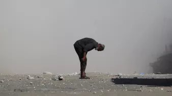 مردی مسن با کمر خمیده در مکانی خاکی و غبارآلود ایستاده است. اطرافش صحنهای ویران وجود دارد.