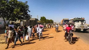 در این تصویر، یک خیابان شنی و پرجنبوجوش دیده میشود. در سمت چپ، تعدادی درخت و ساختمانهای ساده قرار دارند. گروهی از مردم در حال پیادهروی هستند و برخی از آنها کیسههایی به دوش دارند. در سمت راست، چندین خودرو و موتورسیکلت در حال تردد هستند. فضای عمومی پر از فعالیت و زندگی به نظر میرسد.