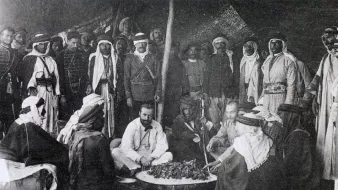 این تصویر تاریخی گروهی از مردان را در یک محیط سنتی نشان میدهد. آنها در یک چادر یا فضای باز گرد هم آمدهاند و به نظر میرسد در حال تهیه یا صرف غذا هستند. چند نفر از آنها لباسهای سنتی عربی به تن دارند، از جمله عبا و headscarf (کشیه). در مرکز تصویر، چند نفر در حال نشستن و گفتگو هستند و سایرین ایستاده یا نشسته در اطراف آنها قرار دارند. فضای تصویر گرم و صمیمی به نظر میرسد و نمایانگر فرهنگی غنی و سنتهای اجتماعی است.