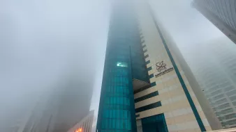 در این تصویر، یک ساختمان بلند و مدرن دیده میشود که در میان مه غلیظ قرار گرفته است. قسمت بالایی ساختمان به طور کامل در مه پنهان شده و جزئیات آن به وضوح دیده نمیشوند. نمای ساختمان شامل شیشههای آبی و رنگهای ملایم دیگر است. در اطراف ساختمان، سایر ساختمانها نیز وجود دارند که بخشی از آنها در مه محو شده است. این صحنه حس اسرارآمیز و آرامشبخشی ایجاد میکند.