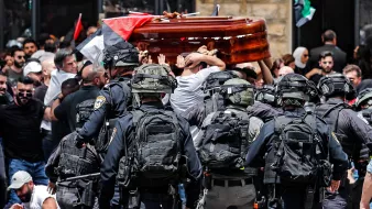 در این تصویر، گروهی از مردم در حال حمل یک تابوت هستند و در اطراف آنها تعدادی نیروی پلیس با تجهیزات نظامی دیده میشود. مردم در حال تجمع و اعتراض هستند و پرچمهایی را در دست دارند. به نظر میرسد این صحنه در یک فضای پرازدحام و تنشزای اجتماعی اتفاق میافتد. واکنشهای متفاوتی در چهرهها و حرکات افراد مشهود است.