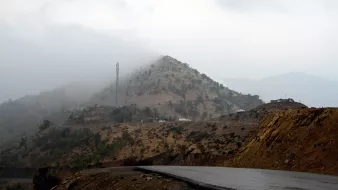در این تصویر، یک جادهای در کوهستان با شیب ملایم دیده میشود. در سمت راست، جاده به سمت دورتر ادامه پیدا میکند و در بالای تپهها، ابرها در حال پایین آمدن و محو کردن قلهها هستند. روی تپهها، درختان نسبتاً کوچک و فضاهای خشک دیده میشوند. در بالای تپه، یک دکل تلفن همراه نیز به چشم میخورد و چندین خودروی سفید رنگ در دور دست قابل مشاهدهاند. جو تصویر ابری و مرطوب به نظر میرسد.