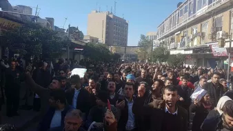در این تصویر، جمعیتی بزرگ در یک خیابان شهری دیده میشود. افراد در حال راهپیمایی هستند و برخی از آنها دستها را بالا برده یا نمادهایی را در دست دارند. ساختمانها و تابلوهای مغازهها در اطراف قابل مشاهدهاند، و به نظر میرسد که این تجمع به منظور ابراز نظر یا اعتراض صورت گرفته است. آسمان روشن بوده و در پسزمینه، سازههای بلند دیده میشوند. جو عمومی تصویر نشاندهنده حرکت و فعالیت جمعیت است.