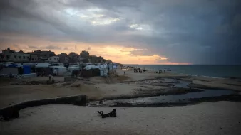 تصویر نشاندهندهی یک ساحل است که در آن آسمان پهناوری با ابرهای تیره و روشن به تصویر کشیده شده است. خورشید در حال غروب است و نور ملایمی بر روی آب دریا و شنهای ساحل میتابد. در سمت چپ تصویر، برخی چادرها و سازههایی دیده میشود که نشاندهندهی سکونتگاههای موقت هستند. همچنین، در دوردست، افرادی در حال حرکت و فعالیت هستند. این تصویر حال و هوای خاصی از مکانهای ساحلی همراه با چالشهای اجتماعی و محیطی را منتقل میکند.