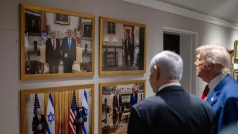 دو مرد در کت و شلوار در کنار یکدیگر ایستادهاند. تصاویر آنها بر دیواری قاب شدهاند.