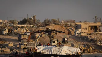 در این تصویر، یک صحنه ساختمانی در کنار دریا دیده میشود. در ساحل، تعدادی ماشین سنگین و جرثقیل در حال کار هستند. یک قایق روی آب قرار دارد که زیر آن یک پوشش سفید رنگ قرار دارد. پسزمینه شامل ساختمانهای نیمهکار و تخریب شده و چند درخت نخل است. نور خورشید به صورت گرم بر روی این صحنه تابیده و حال و هوای آن را به تصویر کشیده است.